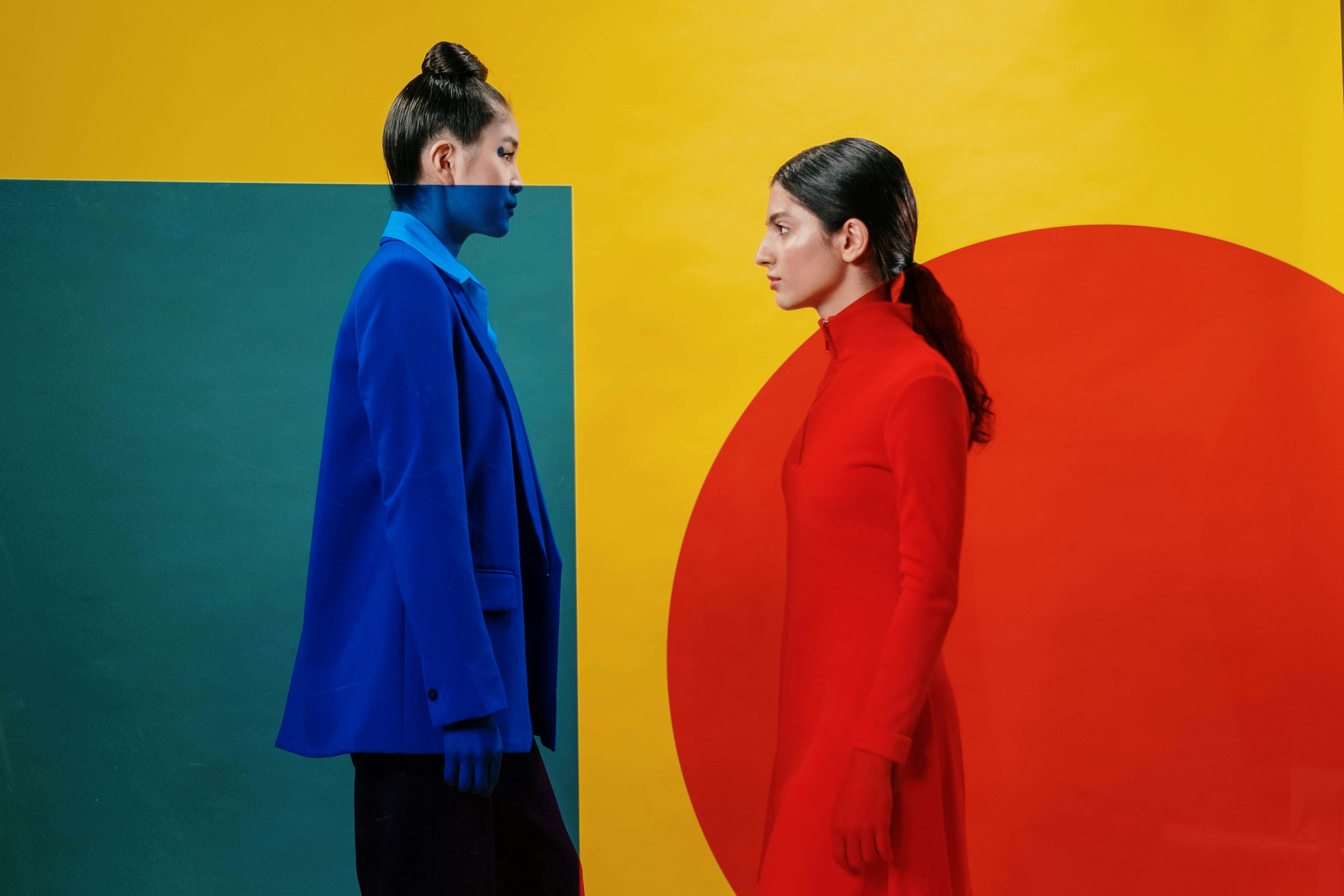 Artistic studio portrait of two women dressed in vibrant red and blue against a geometric background.